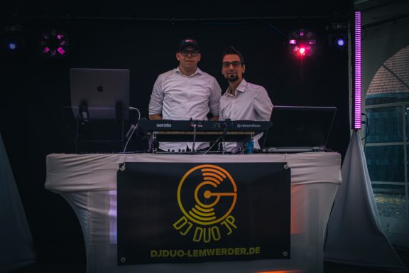 Zwei DJs stehen hinter einem Mixer vor einem beleuchteten Hintergrund.
