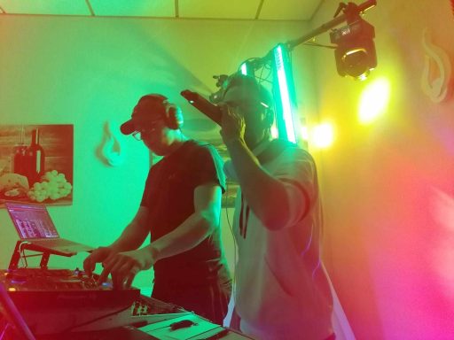 Zwei Männer performen live, einer singt, der andere am DJ-Pult mit bunten Lichtern im Hintergrund.